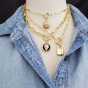 May include: A layered gold chain necklace with three different pendant charms. The top charm is a sunburst with a clear rhinestone in the center. The middle charm is a round medallion with a black and gold lion's head design. The bottom charm is a gold padlock.