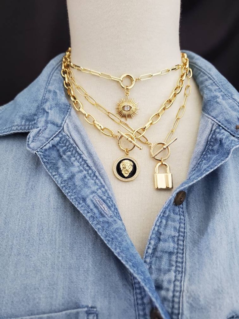 May include: A layered gold chain necklace with three different pendant charms. The top charm is a sunburst with a clear rhinestone in the center. The middle charm is a round medallion with a black and gold lion's head design. The bottom charm is a gold padlock.