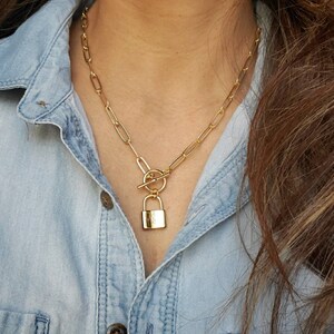May include: A gold chain necklace with a padlock pendant. The chain is made of linked ovals and has a toggle clasp.