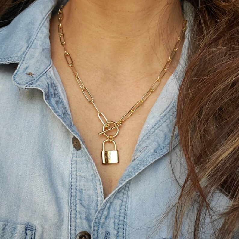 May include: A gold chain necklace with a padlock pendant. The chain is made of linked ovals and has a toggle clasp.