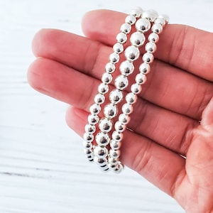 May include: Three silver bead bracelets stacked on top of each other. The bracelets are made of small, round beads.