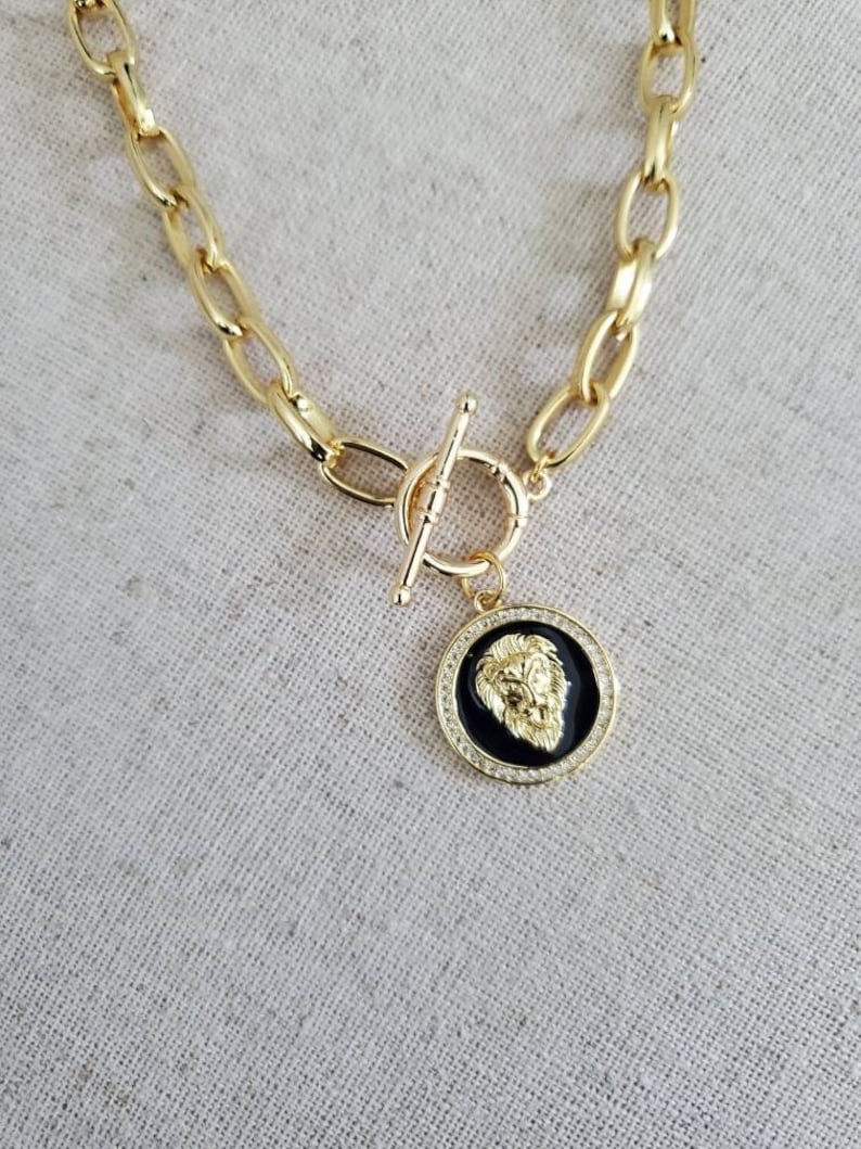 May include: A gold chain necklace with a toggle clasp and a round pendant featuring a lion head design with black enamel and clear rhinestones.
