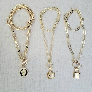 May include: Three gold chain necklaces with different pendants. The first necklace has a lion head pendant. The second necklace has a sun with an evil eye pendant. The third necklace has a padlock pendant.