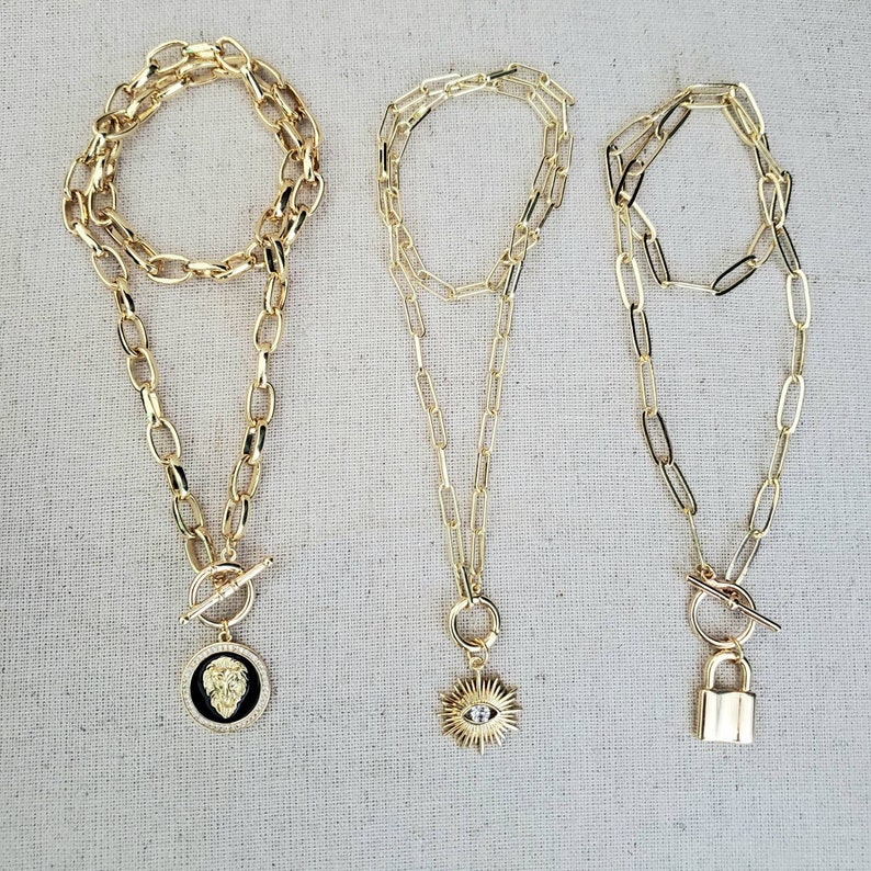 May include: Three gold chain necklaces with different pendants. The first necklace has a lion head pendant. The second necklace has a sun with an evil eye pendant. The third necklace has a padlock pendant.