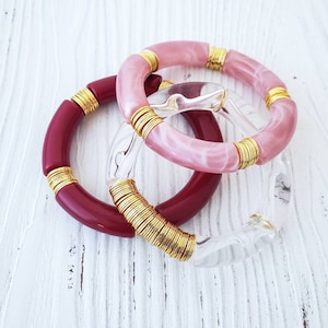 May include: Three bangle bracelets with gold accents. One is pink with white swirls, one is burgundy, and one is clear.