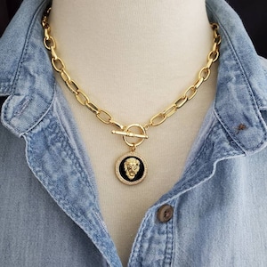 May include: A gold chain necklace with a large, round pendant featuring a lion's head in black enamel. The chain is made of thick, oval-shaped links.