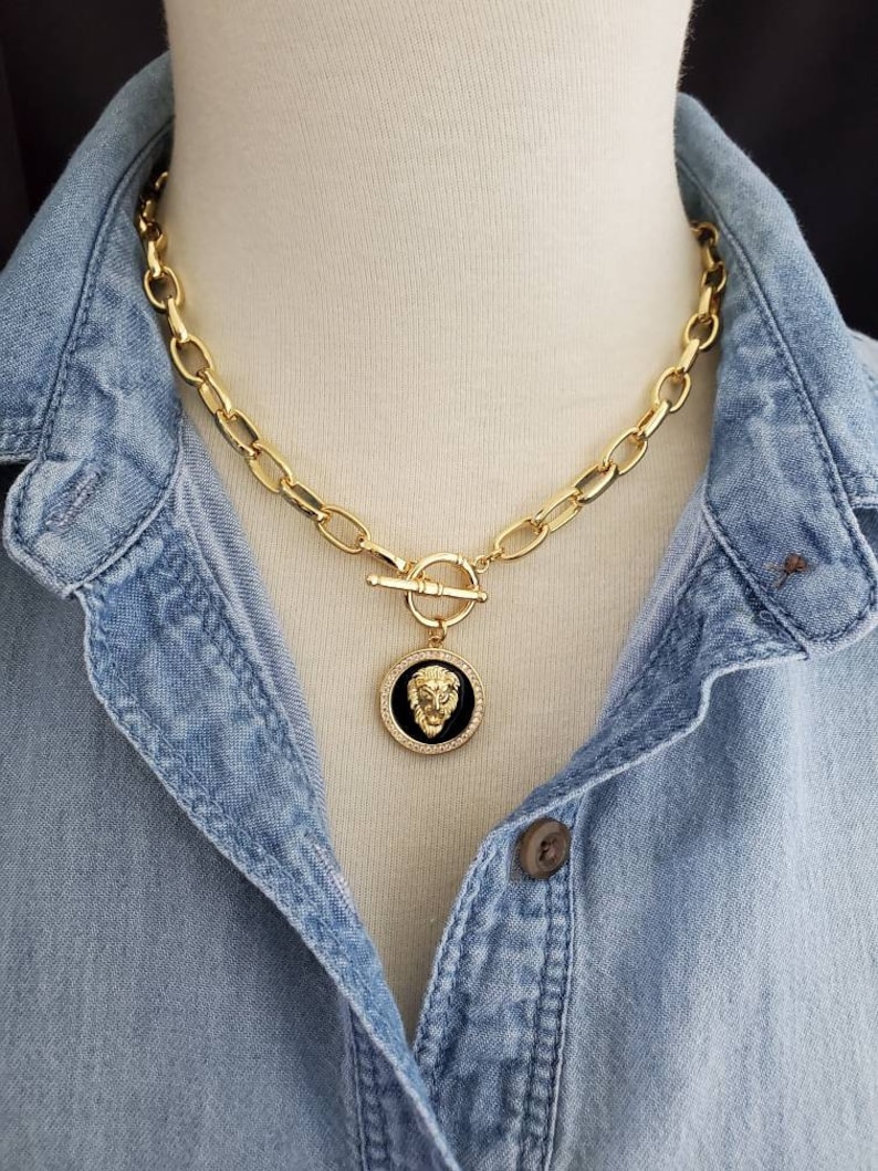 May include: A gold chain necklace with a large, round pendant featuring a lion's head in black enamel. The chain is made of thick, oval-shaped links.