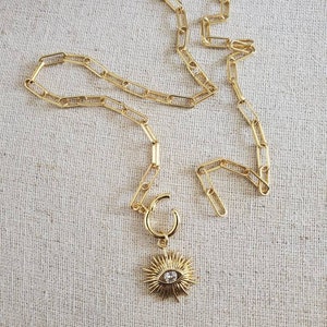 May include: A gold chain necklace with a gold pendant featuring a sunburst design with a clear crystal eye in the center.