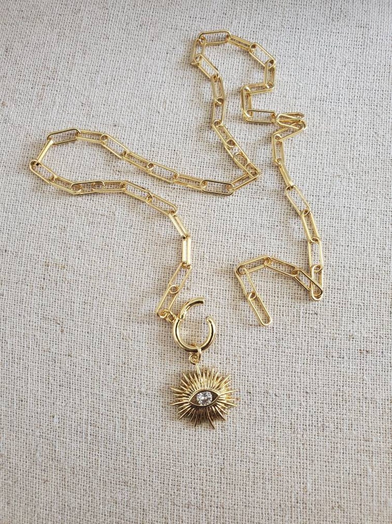 May include: A gold chain necklace with a gold pendant featuring a sunburst design with a clear crystal eye in the center.