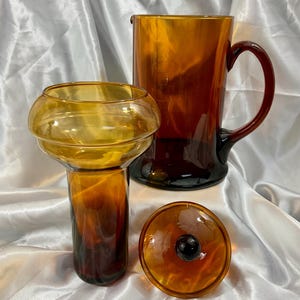May include: A set of amber-colored glassware consisting of a large pitcher with a curved handle, a tall vase with a wide, rounded upper section, and a circular lid featuring a small, dark brown spherical knob at the center of the piece.
