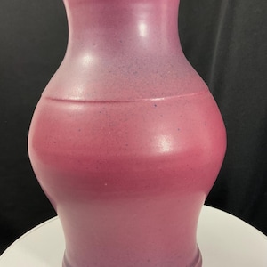 May include: A ceramic vase with a bulbous body and a flared rim, featuring a matte finish in shades of magenta and deep purple. The surface has a subtle speckled texture throughout, with a smooth transition between the pink and purple tones.
