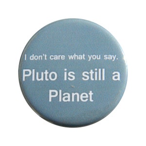 May include: A circular blue pinback button featuring the white text "I don't care what you say. Pluto is still a Planet" printed in a sans-serif font. The button has a smooth, glossy finish and a simple, clean design centered on the face of the object.