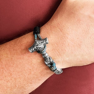 May include: A silver-colored Thor's hammer bracelet with intricate detailing. The hammer is attached to a black and teal braided cord. The bracelet is worn on a wrist against a maroon background.