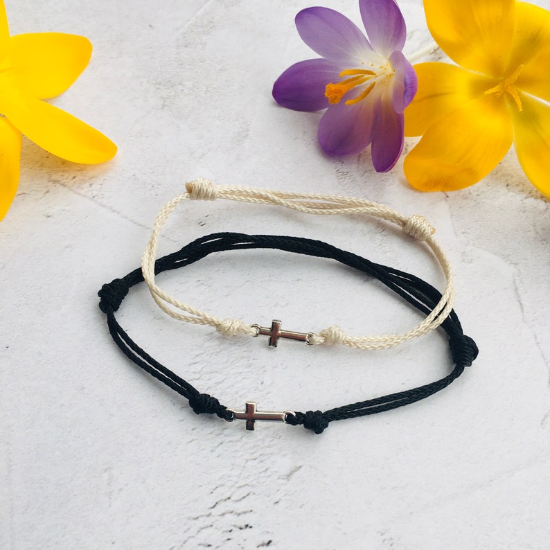 May include: Two adjustable cord bracelets, one black and one cream, each featuring a small silver cross charm. The bracelets are knotted for adjustability and are displayed on a white surface with yellow and purple flowers.