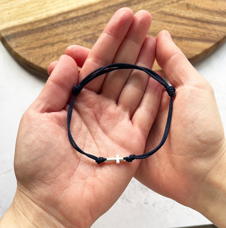 May include: A navy blue cord bracelet with a small silver cross charm. The bracelet is adjustable and features a sliding knot closure. The cross is centered on the bracelet. The bracelet is shown against a light background.