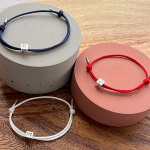 May include: Three adjustable bracelets with silver cube beads. One bracelet is navy blue, one is red, and one is white. Each cube bead has a letter on it. The bracelets are displayed on gray and coral-colored pedestals.
