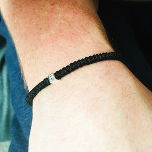 Könnte beinhalten: Schwarz geflochtenes Armband mit einer kleinen quadratischen, silberfarbenen Perle. Das Armband wird am Handgelenk getragen und scheint aus einem strapazierfähigen, gewebten Material zu bestehen. Die Perle hat einen Buchstaben.