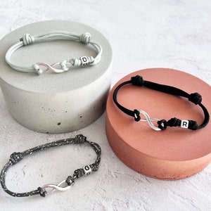 Könnte beinhalten: Drei verstellbare Armbänder mit Unendlichkeitszeichen. Eines ist hellgrau mit dem Buchstaben "D", ein anderes ist schwarz mit dem Buchstaben "R", und das dritte ist grau und schwarz mit dem Buchstaben "G". Jedes Armband hat einen silbernen Unendlichkeits-Anhänger.