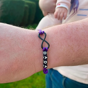 May include: A black infinity symbol bracelet with the word "DADA" spelled out in white letter beads. The bracelet is accented with purple cord. The bracelet is worn on a wrist.