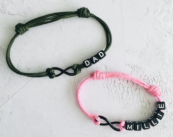 Puede incluir: Dos pulseras ajustables con símbolos de infinito. Una es verde oliva con la palabra "DAD" en cuentas de letras negras. La otra es rosa con el nombre "MILLIE" en cuentas de letras negras. Ambas pulseras tienen un diseño anudado.