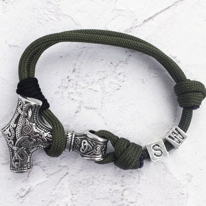 Puede incluir: Pulsera de cordón verde y negro con un dije de martillo de Thor plateado y un dije cilíndrico. La pulsera incluye cuentas de letras plateadas que deletrean "S W". Cierre con nudo.