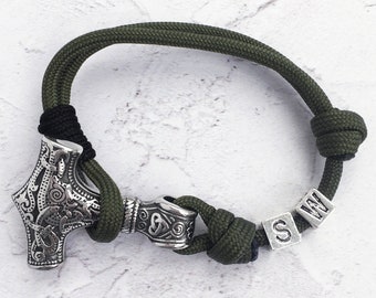 Puede incluir: Pulsera de cordón verde y negro con un dije de martillo de Thor plateado y un dije cilíndrico. La pulsera incluye cuentas de letras plateadas que deletrean "S W". Cierre con nudo.