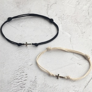 May include: Two adjustable bracelets with a cross charm. One bracelet is black, the other is off-white. Each bracelet features a small silver-colored cross pendant and knotted accents. The bracelets are displayed on a light-colored surface.