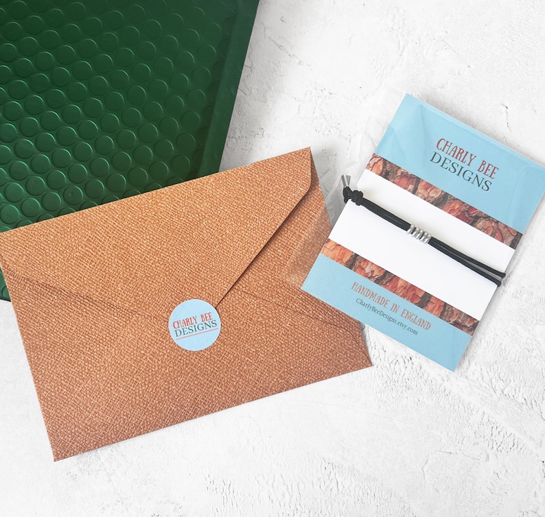 K&ouml;nnte beinhalten: Draufsicht auf einen braunen Umschlag mit einem blauen Aufkleber "Charly Bee Designs". Ein gr&uuml;ner Luftpolsterumschlag und eine blaue Karte mit einem schwarzen Armband sind ebenfalls zu sehen. Auf der Karte steht "Charly Bee Designs" und "Handmade in England".