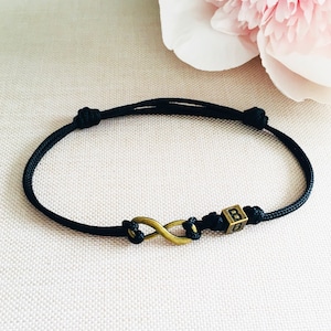 Può includere: Braccialetto con cordino nero con un simbolo infinito color bronzo e un piccolo cubo con la lettera "B". Il braccialetto è regolabile con nodi.