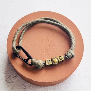 May include: A khaki-colored paracord bracelet with a black carabiner clasp. The bracelet features three gold-colored letter beads spelling out "PGC" with a heart symbol. The bracelet is displayed on a round, reddish-brown surface.