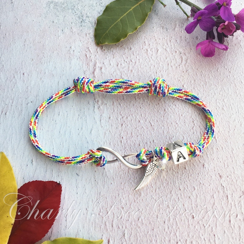 K&ouml;nnte beinhalten: Ein mehrfarbiges Freundschaftsarmband mit einem Unendlichkeitssymbol und einem silbernen Fl&uuml;gelanh&auml;nger. Das Armband hat einen kleinen W&uuml;rfel mit dem Buchstaben "A". Das Armband besteht aus geflochtenem Kordel und wird auf einer hellen Oberfl&auml;che pr&auml;sentiert.