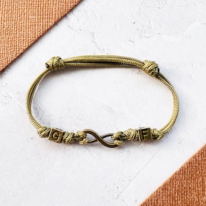 May include: Olive green rope bracelet featuring an infinity symbol and two lettered cubes. The bracelet is knotted at each end. The letters "G" and "F" are visible on the cubes. The bracelet is on a white surface.