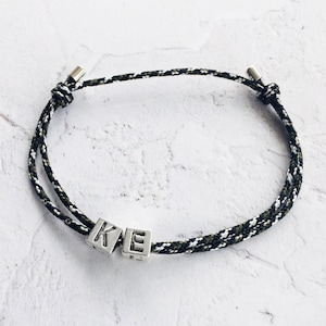 Puede incluir: Una pulsera de cord&oacute;n trenzado negro, blanco y verde oliva, con detalles met&aacute;licos plateados. La pulsera presenta dos cubos plateados con las letras "K" y "E". Se muestra sobre una superficie blanca texturizada.