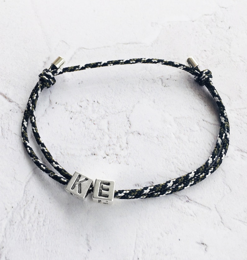 Puede incluir: Una pulsera de cord&oacute;n trenzado negro, blanco y verde oliva, con detalles met&aacute;licos plateados. La pulsera presenta dos cubos plateados con las letras "K" y "E". Se muestra sobre una superficie blanca texturizada.
