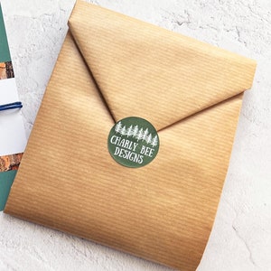 May include: A package wrapped in brown paper with a green circular sticker. The sticker features a white tree line and the text "Charly Bee Designs". The paper is folded and sealed.