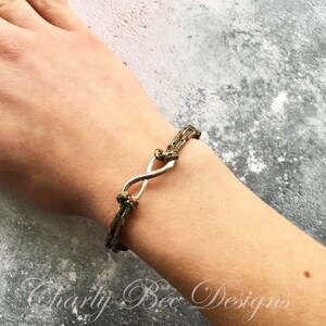 K&ouml;nnte beinhalten: Ein Armband mit Camouflage-Muster und einem silbernen Unendlichkeitszeichen. Das Armband besteht aus mehreren Kordelstr&auml;ngen, die an jedem Ende des Unendlichkeitszeichens verknotet sind. Das Armband befindet sich an einem Handgelenk.