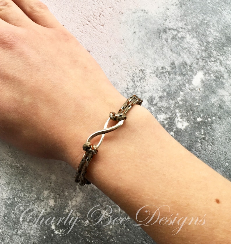 K&ouml;nnte beinhalten: Ein Armband mit Camouflage-Muster und einem silbernen Unendlichkeitszeichen. Das Armband besteht aus mehreren Kordelstr&auml;ngen, die an jedem Ende des Unendlichkeitszeichens verknotet sind. Das Armband befindet sich an einem Handgelenk.