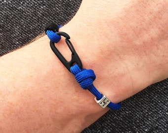 Puede incluir: Una pulsera azul y negra con un cierre de mosquetón negro. La pulsera está hecha de cuerda azul y tiene una pequeña cuenta cuadrada de color plateado. La pulsera se muestra en la muñeca de una persona.