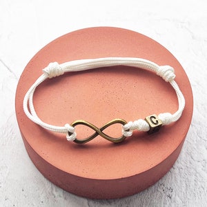 May include: White cord bracelet featuring a bronze-colored infinity symbol and a small cube bead with the letter "C". The bracelet is displayed on a round, coral-colored surface.