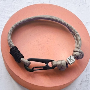 Pode incluir: Uma pulseira de corda cinza e preta com um fecho preto e um berloque inicial prateado. A pulseira está sobre uma superfície circular rosa. O berloque inicial tem a letra "K".