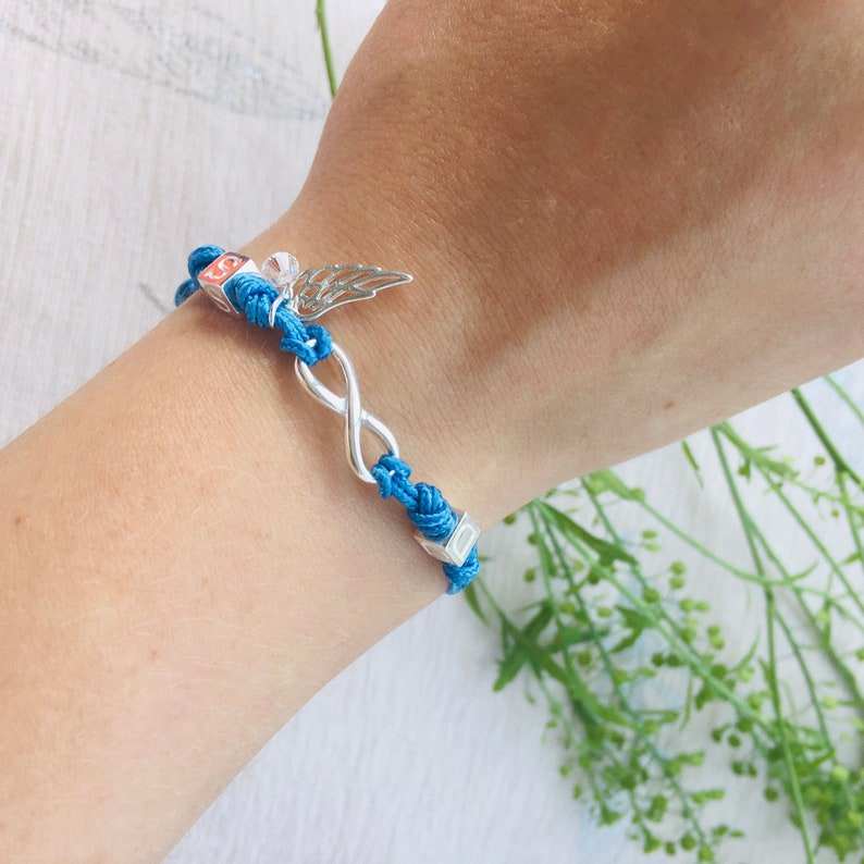 May include: A blue cord bracelet with a silver infinity symbol and a small silver wing charm. The bracelet also features small square beads. The bracelet is on a wrist.