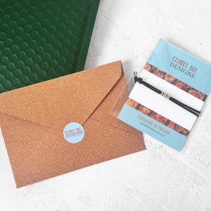 May include: An overhead shot of a brown textured envelope with a blue sticker that says "Charly Bee Designs". A green bubble mailer is in the background. A blue card with a black bracelet and the text "Charly Bee Designs" is also visible.