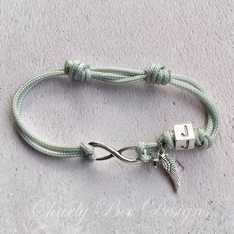 K&ouml;nnte beinhalten: Hellgr&uuml;nes, verstellbares Armband mit Unendlichkeitssymbol und kleinem Fl&uuml;gelanh&auml;nger. Das Armband hat eine W&uuml;rfelperle mit dem Buchstaben "J". Der Text "Charly Bee Designs" ist sichtbar.