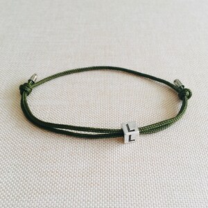 Puede incluir: Una pulsera de cord&oacute;n verde oliva con un peque&ntilde;o dije de cubo plateado. El cubo tiene la letra "L" en dos lados. La pulsera es ajustable y tiene extremos met&aacute;licos plateados.
