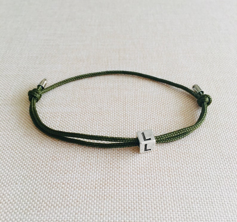 Puede incluir: Una pulsera de cord&oacute;n verde oliva con un peque&ntilde;o dije de cubo plateado. El cubo tiene la letra "L" en dos lados. La pulsera es ajustable y tiene extremos met&aacute;licos plateados.