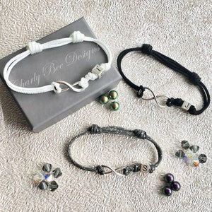May include: Three infinity symbol bracelets in white, black, and grey, each with a knot design. The bracelets have a silver infinity charm and a small initial cube. The bracelets are displayed with decorative beads and a grey box with the text "Charly Bee Designs."