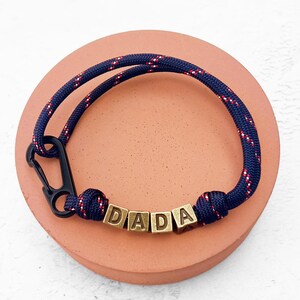 May include: A navy blue rope bracelet with red and white accents and the word "DADA" spelled out in gold-colored letter beads. The bracelet features a black carabiner clasp and is displayed on a round, peach-colored surface.