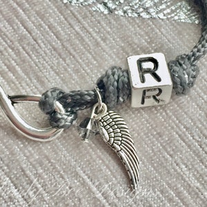 K&ouml;nnte beinhalten: Ein graues Kordelarmband mit einem silbernen Unendlichkeits-Anh&auml;nger und einem kleinen silbernen Fl&uuml;gel-Anh&auml;nger. Das Armband hat einen wei&szlig;en W&uuml;rfel mit dem Buchstaben "R". Das Armband liegt auf einer hellrosa Oberfl&auml;che.