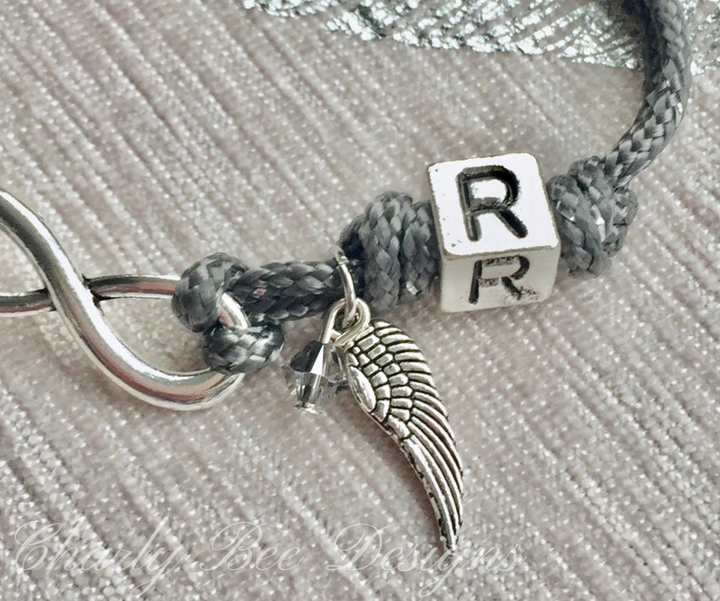 K&ouml;nnte beinhalten: Ein graues Kordelarmband mit einem silbernen Unendlichkeits-Anh&auml;nger und einem kleinen silbernen Fl&uuml;gel-Anh&auml;nger. Das Armband hat einen wei&szlig;en W&uuml;rfel mit dem Buchstaben "R". Das Armband liegt auf einer hellrosa Oberfl&auml;che.