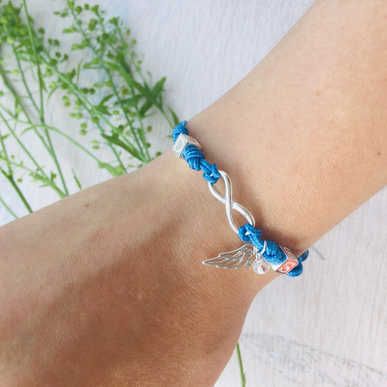 May include: A blue cord bracelet with a silver infinity symbol and a wing charm. The bracelet features small square beads and is knotted. The bracelet is worn on a wrist.